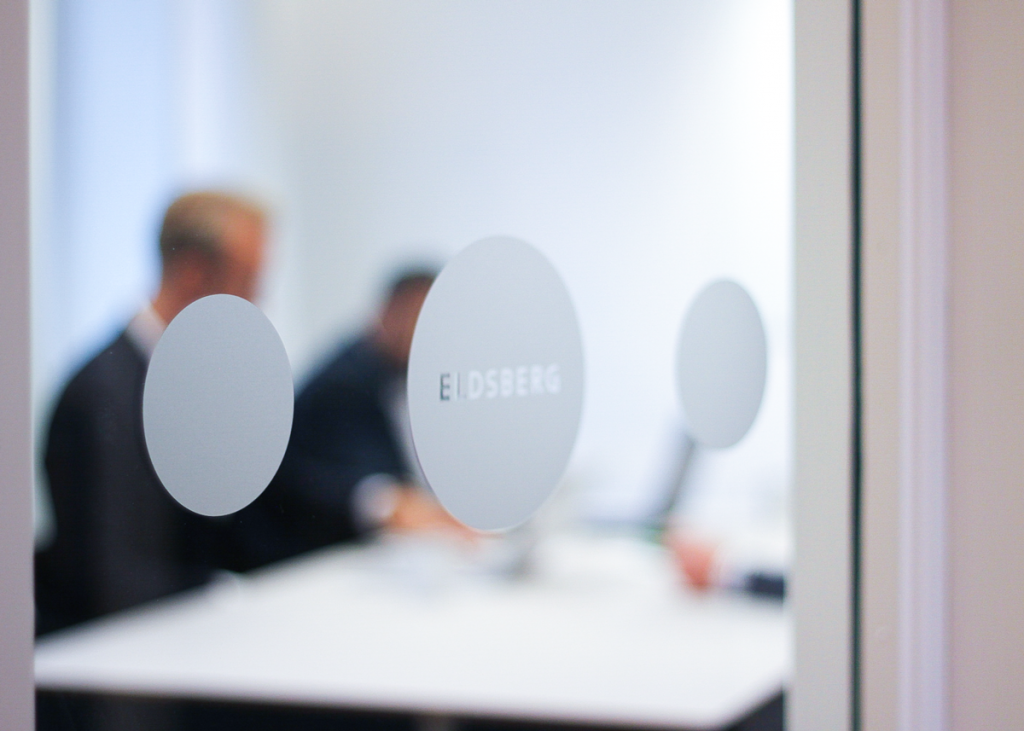 Fotografi på mötesrum med text"ELdsberg" i förgrunden. I bakgrunden ses personer sitta i rummet och arbeta.