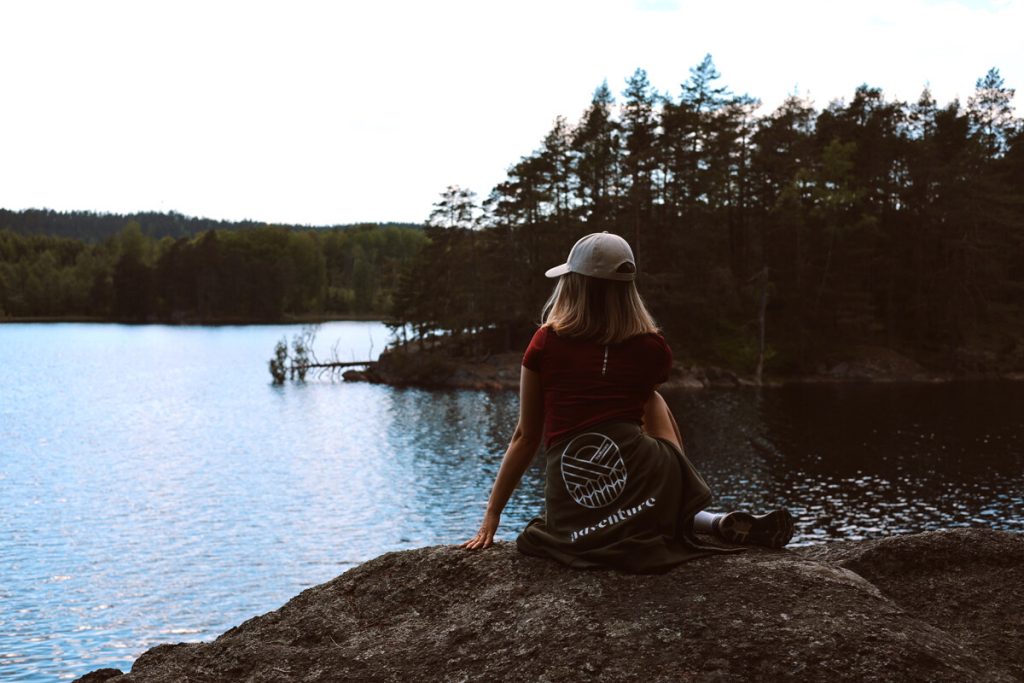 Kvinna med keps och kortklippt hår sitter på en klippavsats och ser ut över sjö